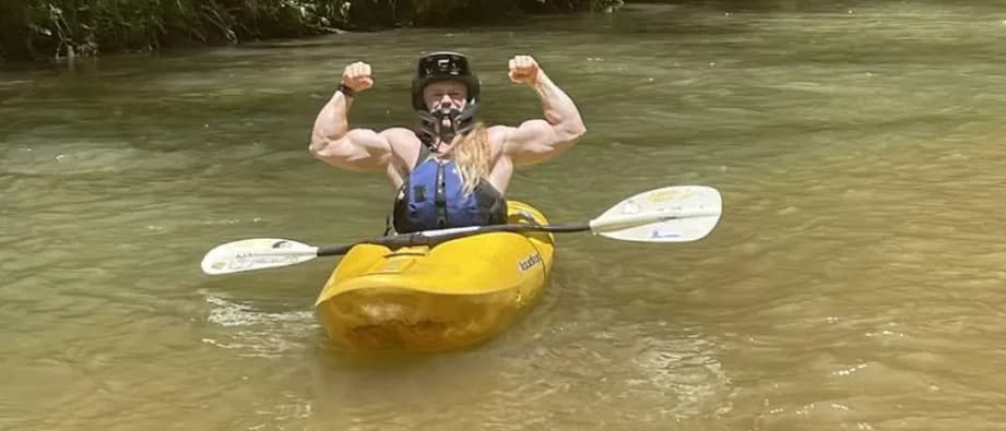 Cody Wood flexing both arms in a yellow whitewater kayak on a calm river